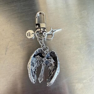 May include: Silver-toned keychain featuring an angel wing design with a cross in the center. The keychain includes a lobster clasp, a star-shaped charm, and a small round charm with the text "LOVE YOU".