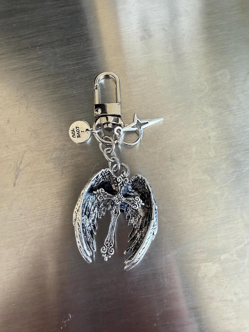 May include: Silver-toned keychain featuring an angel wing design with a cross in the center. The keychain includes a lobster clasp, a star-shaped charm, and a small round charm with the text "LOVE YOU".