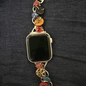 May include: Silver Apple Watch with a colourful button band. The band features a variety of round buttons in red, black, yellow, and blue, connected by silver links. The watch face is black.