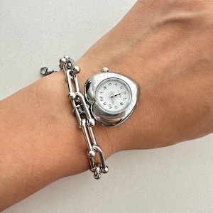 Könnte beinhalten: Eine silberne herzförmige Uhr mit weißem Zifferblatt und schwarzen Zahlen. Die Uhr ist an einem silbernen Gliederarmband befestigt.