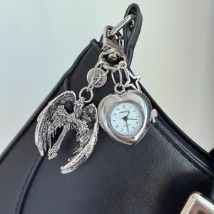 May include: Silver-toned keychain with a heart-shaped watch, angel wings with a cross, and star charm. The watch has a white face with black numerals and hands. The keychain is attached to a black bag.