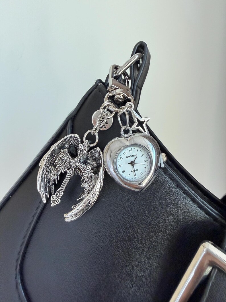 May include: Silver-toned keychain with a heart-shaped watch, angel wings with a cross, and star charm. The watch has a white face with black numerals and hands. The keychain is attached to a black bag.