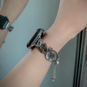 May include: Silver-toned Apple Watch band with a decorative design featuring circular embellishments and a dangling charm. The band is attached to a black Apple Watch.