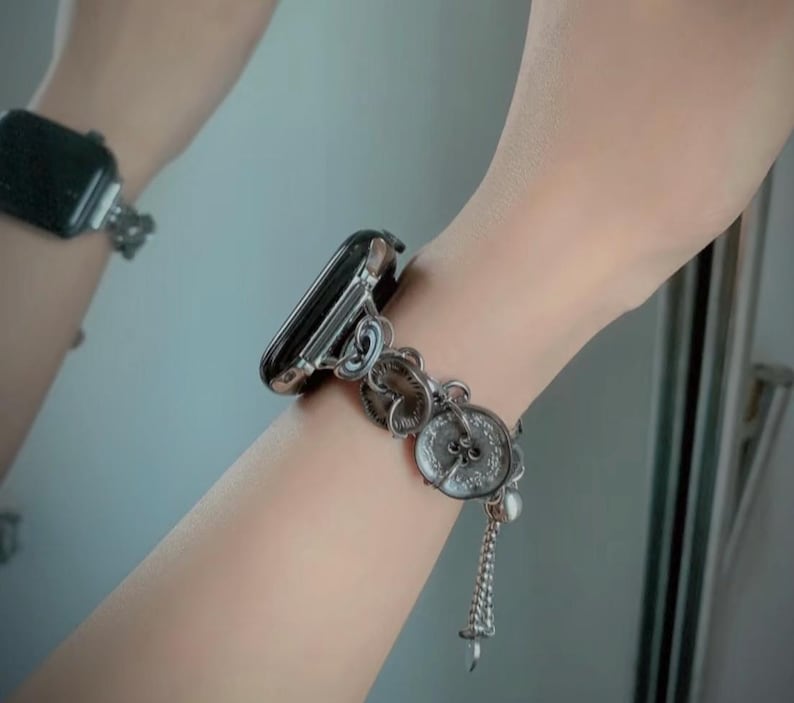 May include: Silver-toned Apple Watch band with a decorative design featuring circular embellishments and a dangling charm. The band is attached to a black Apple Watch.