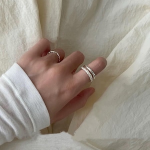 May include: A hand wearing a white long-sleeved shirt displays three silver rings. One ring has a textured design, while the other two are stacked, with one featuring small, clear stones. The background is a soft, off-white fabric.