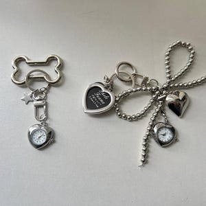 Może przedstawiać: Dwa srebrne breloki do kluczy z zawieszkami. Jeden brelok ma pierścień w kształcie kości z gwiazdą i mały zegarek z srebrną kopertą w kształcie serca. Drugi brelok ma srebrną ramkę do zdjęć w kształcie serca z napisem "Photo Frame" i srebrny zegarek w kształcie serca z srebrnym łańcuszkiem.