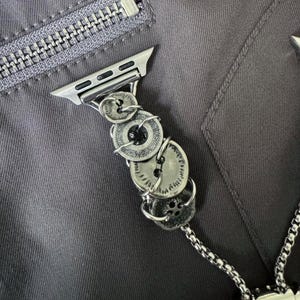 May include: Silver-toned Apple Watch band featuring a unique design of stacked, vintage-style buttons. The buttons are connected with wire and attached to a silver watch adapter. The band is shown against a gray fabric background.