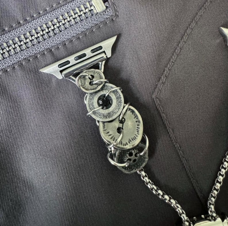May include: Silver-toned Apple Watch band featuring a unique design of stacked, vintage-style buttons. The buttons are connected with wire and attached to a silver watch adapter. The band is shown against a gray fabric background.