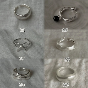 May include: A collection of silver rings in various designs. One ring has a textured surface, another features a bow, and a third has a black bead. Each ring is displayed on a neutral fabric background. Numbers 25, 26, 27, 28, 29, and 30 are visible.