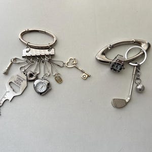Puede incluir: Dos llaveros plateados. El primer llavero tiene un abridor de botellas, un reloj pequeño, un dije de corazón y un dije de llave. El segundo llavero tiene un dije de palo de golf y un dije de pelota de golf. Ambos llaveros tienen el texto "Safety First Drink With A Nurse".