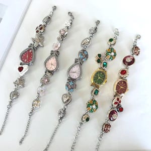 May include: Five silver-tone charm bracelet watches arranged vertically. Each watch features a unique dial color including red, light pink, and green, accented with various charms such as hearts, flowers, stars, and colorful gemstones along the chain.
