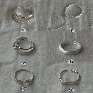 May include: A collection of silver rings, each with a unique design. The rings include adjustable styles, textured bands, and open designs. The image shows rings labeled 13, 14, 15, 16, 17, and 18. The rings are displayed on a neutral fabric background.