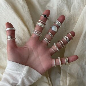 May include: A hand displaying a collection of silver rings. The rings vary in design, including bands, textured styles, and rings with gemstones and pearls. The rings are worn on multiple fingers, showcasing a variety of styles and sizes.