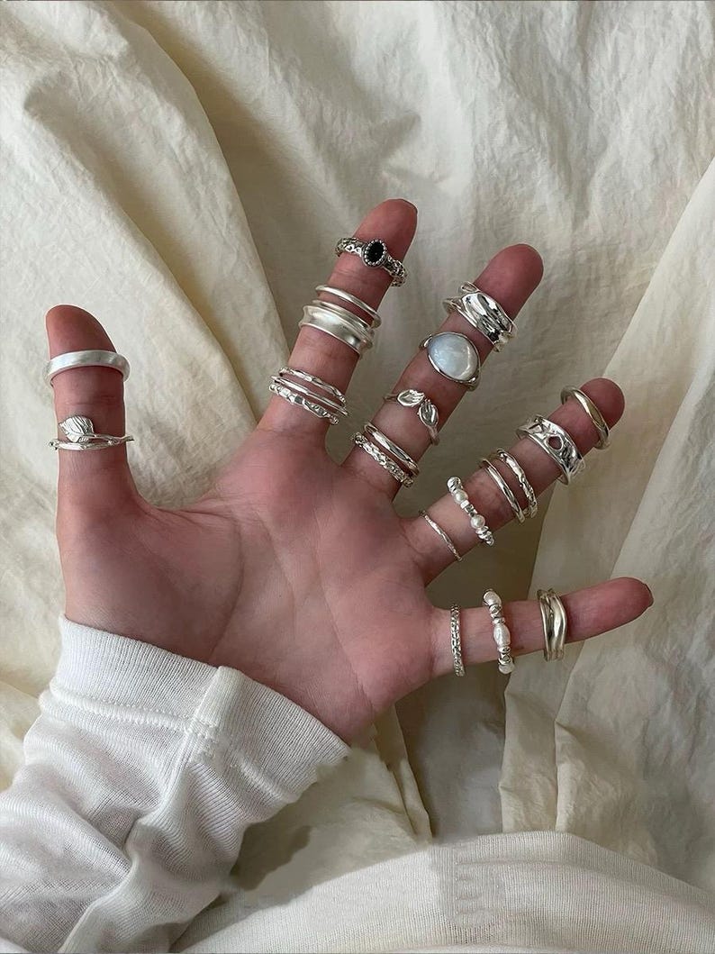 May include: A hand displaying a collection of silver rings. The rings vary in design, including bands, textured styles, and rings with gemstones and pearls. The rings are worn on multiple fingers, showcasing a variety of styles and sizes.