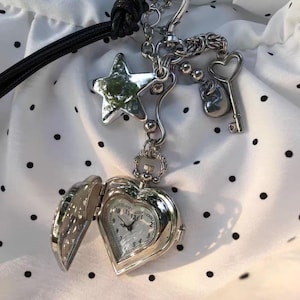 May include: A silver-toned heart-shaped locket watch with an open face, revealing a clock face. The watch is attached to a chain with charms, including a star, a key, and a small round charm. The watch is on a black cord.