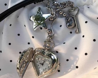 Könnte beinhalten: Eine silberfarbene herzförmige Taschenuhr mit offenem Zifferblatt. Die Uhr ist an einer Kette mit Anhängern befestigt, darunter ein Stern, ein Schlüssel und ein kleiner runder Anhänger. Die Uhr befindet sich an einem schwarzen Band.