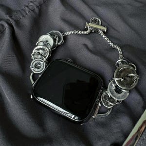 May include: A silver-toned watch band with a unique design. The band features a series of decorative, circular elements, possibly buttons, connected by a delicate chain. The watch face is black and rectangular, contrasting with the metallic band. The watch is resting on a dark gray surface.