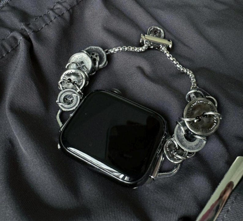 May include: A silver-toned watch band with a unique design. The band features a series of decorative, circular elements, possibly buttons, connected by a delicate chain. The watch face is black and rectangular, contrasting with the metallic band. The watch is resting on a dark gray surface.