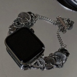 May include: A silver-toned watch band with a series of decorative, circular elements. The band is designed to fit a smartwatch, with a rectangular black screen visible. The band features a chain-like design and a clasp closure.