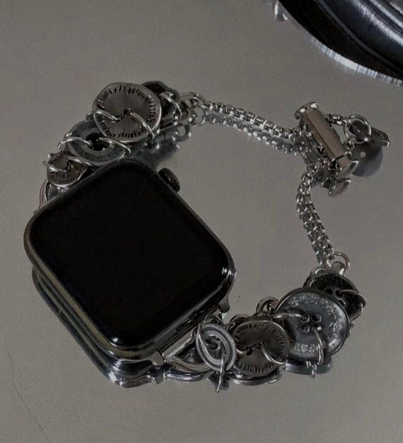 May include: A silver-toned watch band with a series of decorative, circular elements. The band is designed to fit a smartwatch, with a rectangular black screen visible. The band features a chain-like design and a clasp closure.