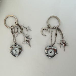 K&ouml;nnte beinhalten: Zwei silberne Schl&uuml;sselanh&auml;nger mit herzf&ouml;rmigen Uhren. Jeder Schl&uuml;sselanh&auml;nger hat eine Kette und himmlische Charms, darunter Sterne und ein Mond. Die Uhren haben wei&szlig;e Zifferbl&auml;tter und schwarze Zeiger.