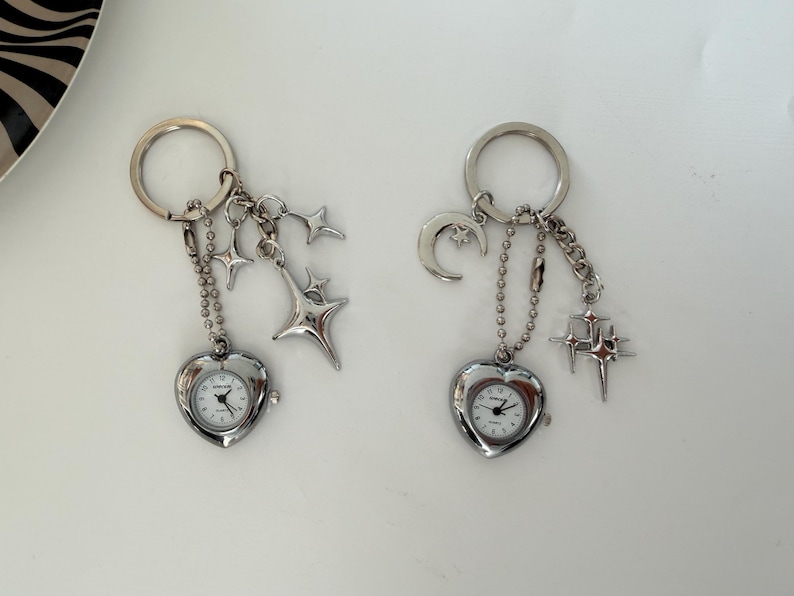 K&ouml;nnte beinhalten: Zwei silberne Schl&uuml;sselanh&auml;nger mit herzf&ouml;rmigen Uhren. Jeder Schl&uuml;sselanh&auml;nger hat eine Kette und himmlische Charms, darunter Sterne und ein Mond. Die Uhren haben wei&szlig;e Zifferbl&auml;tter und schwarze Zeiger.