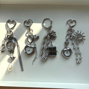Puede incluir: Una colección de llaveros plateados con relojes en miniatura y dijes decorativos. Los llaveros incluyen esferas de reloj en forma de corazón, rectangulares y redondas, con adornos como perlas, rayos de sol y cadenas de cuentas. Cada llavero tiene un cierre en forma de corazón.