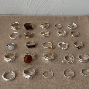 May include: A collection of silver rings with various designs, including bands, gemstone settings, and textured surfaces. Some rings feature natural stones in shades of brown, while others have delicate pearl or bead accents. The rings are displayed on a neutral-toned fabric.