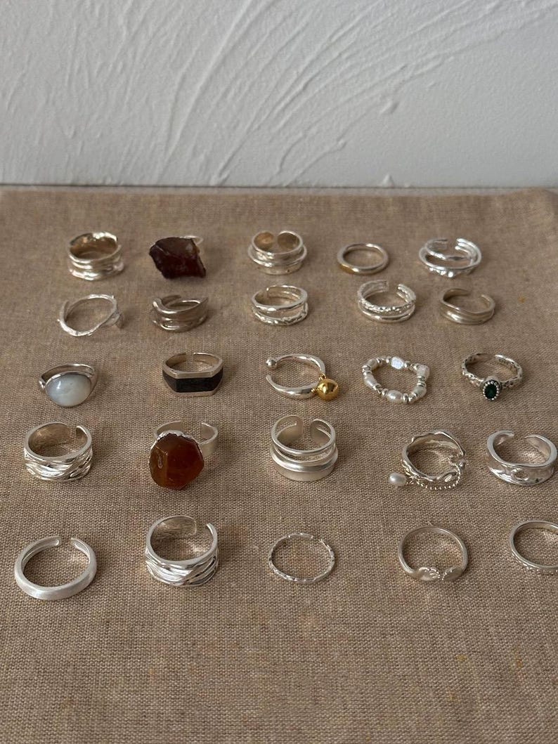 May include: A collection of silver rings with various designs, including bands, gemstone settings, and textured surfaces. Some rings feature natural stones in shades of brown, while others have delicate pearl or bead accents. The rings are displayed on a neutral-toned fabric.
