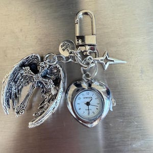May include: A silver-toned keychain featuring a heart-shaped watch, angel wings with a cross, and a star charm. The watch face is white with black numerals and hands. The keychain includes a lobster clasp and a small charm that says "Love You".