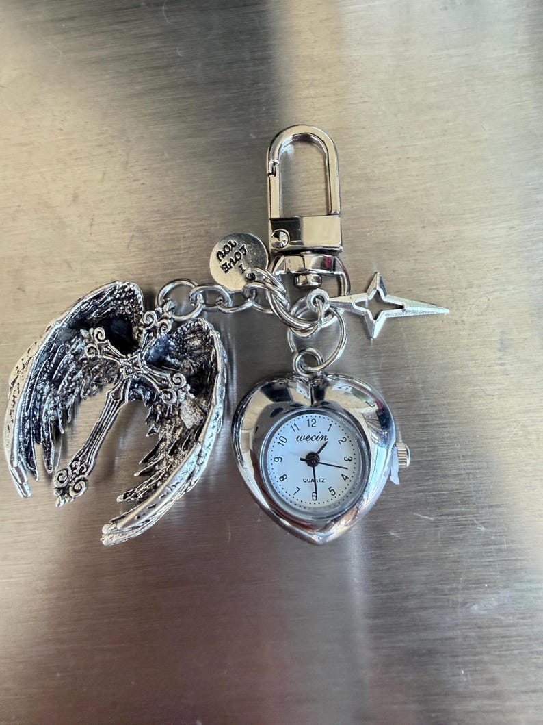 May include: A silver-toned keychain featuring a heart-shaped watch, angel wings with a cross, and a star charm. The watch face is white with black numerals and hands. The keychain includes a lobster clasp and a small charm that says "Love You".