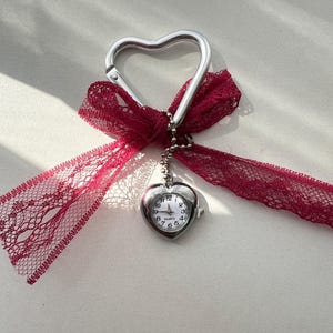 Puede incluir: Un llavero de plata en forma de corazón con un pequeño reloj en forma de corazón adjunto. El llavero tiene un lazo de encaje rojo atado alrededor.