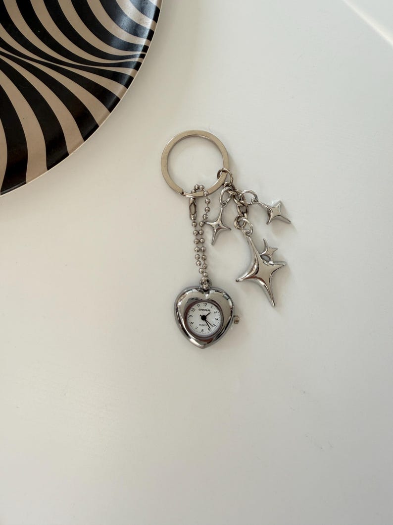 K&ouml;nnte beinhalten: Silberfarbener Schl&uuml;sselanh&auml;nger mit einer herzf&ouml;rmigen Uhr mit wei&szlig;em Zifferblatt und schwarzen Zeigern. Der Schl&uuml;sselanh&auml;nger enth&auml;lt Sternanh&auml;nger und eine Kette. Die Uhr zeigt die Uhrzeit an. Der Hintergrund ist wei&szlig;.