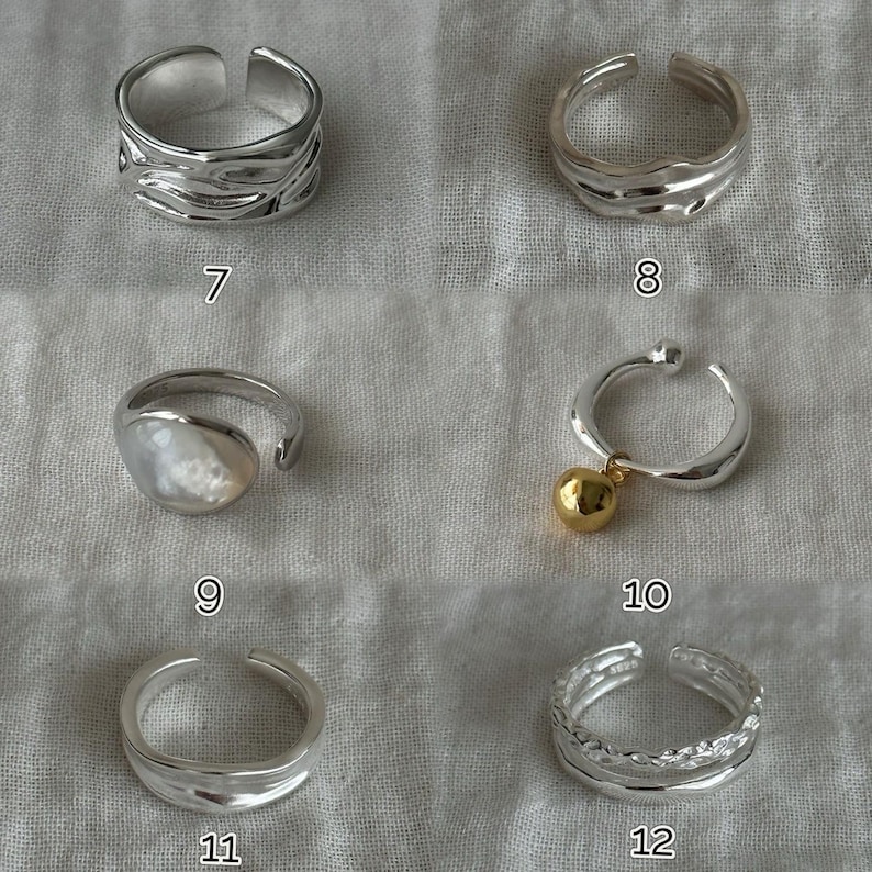 May include: A collection of silver-toned rings. Designs include textured bands, a ring with a pearl-like stone, a ring with a gold-colored ball charm, and a double-band ring with a textured pattern. Each ring is open-ended.