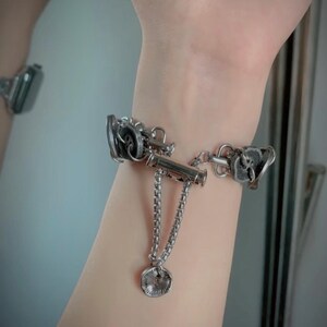 May include: Silver-toned bracelet featuring a unique design with intricate mechanical-style elements. The bracelet includes a central bar with dangling chains and a circular charm. The design is detailed and eye-catching, suitable for a bold fashion statement.