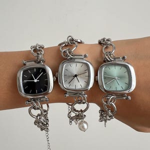May include: Three silver wristwatches with square faces and chain link straps. The watch faces are black, white, and light green. The watch on the right has a pearl charm.