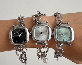 May include: Three silver wristwatches with square faces and chain link bands. The watch faces are black, white, and light green. The watch on the right has a pearl charm.