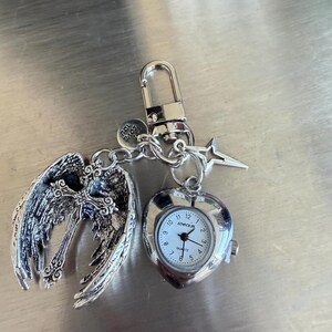 May include: Silver-toned keychain with a heart-shaped watch, angel wings, and a star charm. The watch face is white with black numerals and hands. The angel wings are detailed with intricate feather designs. The keychain includes a lobster clasp and a small charm that says "Love You".