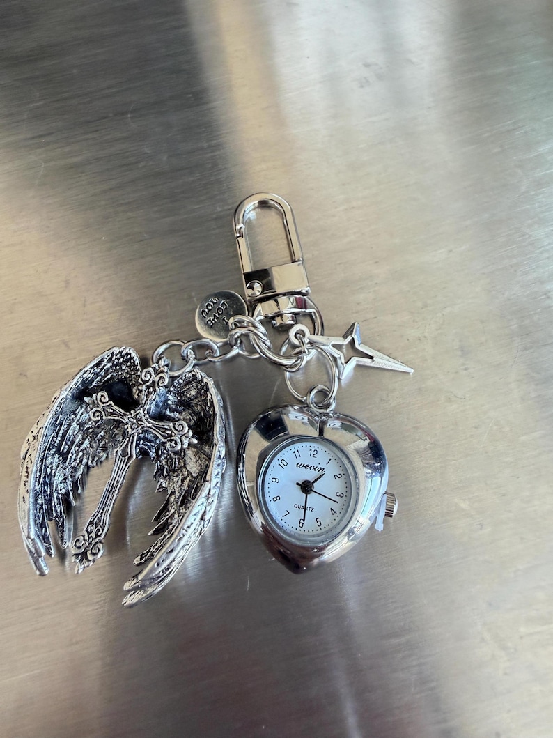 May include: Silver-toned keychain with a heart-shaped watch, angel wings, and a star charm. The watch face is white with black numerals and hands. The angel wings are detailed with intricate feather designs. The keychain includes a lobster clasp and a small charm that says "Love You".
