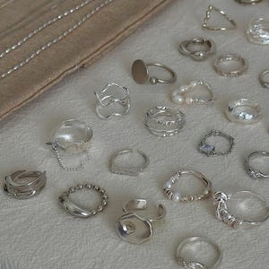 May include: A collection of silver rings in various designs, including bands, geometric shapes, and rings with pearl accents. The rings are displayed on a cream-colored surface, with a neutral-toned fabric in the background.