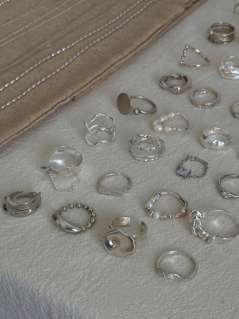May include: A collection of silver rings in various designs, including bands, geometric shapes, and rings with pearl accents. The rings are displayed on a cream-colored surface, with a neutral-toned fabric in the background.