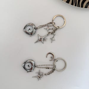 K&ouml;nnte beinhalten: Zwei silberne Schl&uuml;sselanh&auml;nger mit herzf&ouml;rmigen Uhren. Einer hat Sternanh&auml;nger, der andere einen Mond- und Sternanh&auml;nger. Beide haben eine silberne Kette und einen Schl&uuml;sselring.