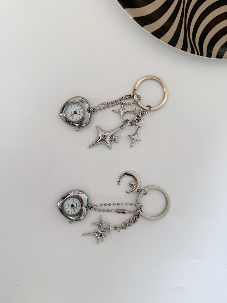 K&ouml;nnte beinhalten: Zwei silberne Schl&uuml;sselanh&auml;nger mit herzf&ouml;rmigen Uhren. Einer hat Sternanh&auml;nger, der andere einen Mond- und Sternanh&auml;nger. Beide haben eine silberne Kette und einen Schl&uuml;sselring.