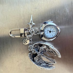 May include: Silver-toned keychain featuring a small pocket watch, a cross pendant with angel wings, and a charm that says "Love You." The watch has a white face with black numbers and hands. The angel wings and cross are detailed.