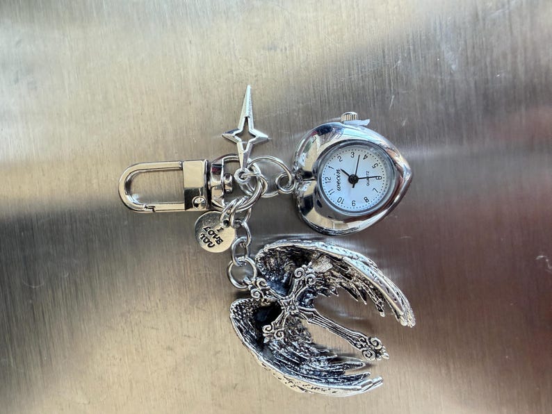 May include: Silver-toned keychain featuring a small pocket watch, a cross pendant with angel wings, and a charm that says "Love You." The watch has a white face with black numbers and hands. The angel wings and cross are detailed.