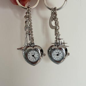 K&ouml;nnte beinhalten: Zwei silberfarbene herzf&ouml;rmige Schl&uuml;sselanh&auml;nger mit kleinen analogen Uhren. Jede Uhr hat ein wei&szlig;es Zifferblatt mit schwarzen Ziffern und Zeigern. Die Schl&uuml;sselanh&auml;nger haben dekorative Anh&auml;nger.
