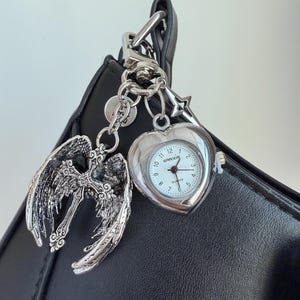 May include: A silver-toned keychain with a heart-shaped watch, angel wings, and a cross charm. The watch face is white with black numerals and hands. The keychain is attached to a black leather bag.