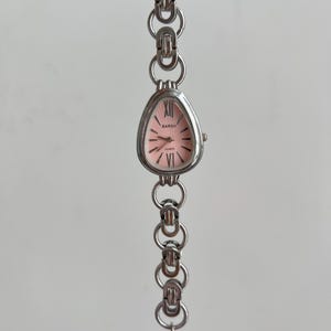 Op de afbeelding: Een zilveren horloge met een roze wijzerplaat en Romeinse cijfers voor de uren. Het horloge heeft een zilveren metalen band met een sluiting.