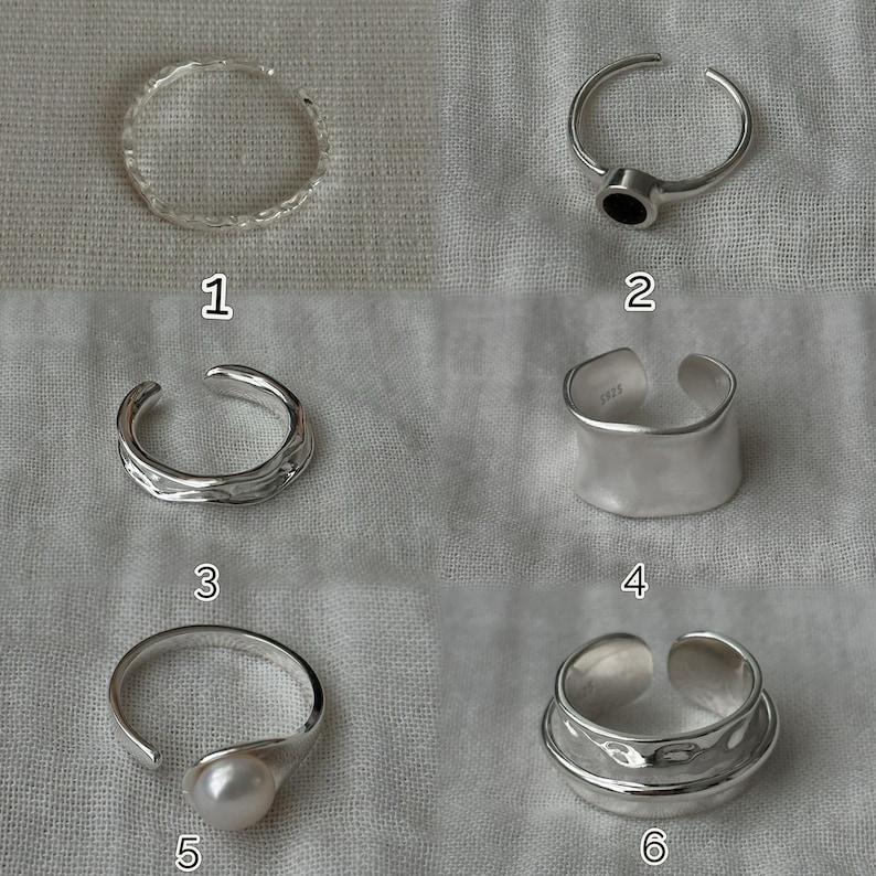 May include: A collection of six silver-toned rings. Designs include a textured band, a ring with a black stone, a hammered band, a ring with a pearl, and a double-banded ring. The rings are displayed on a light-colored fabric.