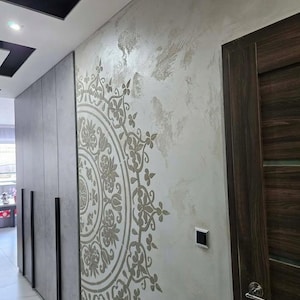 May include: A hallway with a decorative wall featuring a large, circular floral design in shades of gray and beige. The wall has a textured finish. Dark gray cabinets with black handles line one side, and a dark wood door is on the other.